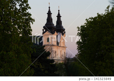 A striking European church with twin spires is framed by vibrant green foliage during the golden hour, creating a picturesque scene 141323849