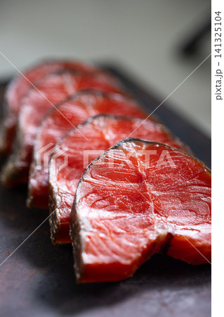 Closeup row of glossy smoked sockeye salmon slices on dark board, rich red flesh, thin darkened edges, visible muscle striations, soft natural light and shallow focus Closeup row of glossy smoked sockeye salmon slices on dark board, rich red flesh, thin darkened edges, visible muscle striations, soft natural light and shallow focus 141325104