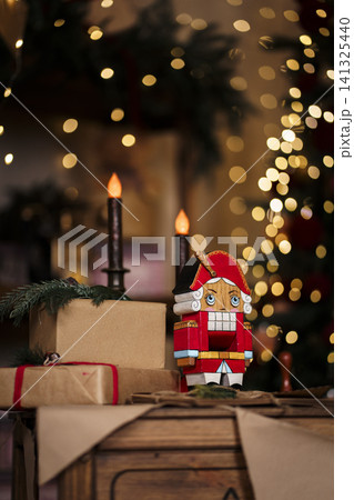 Wooden Nutcracker near gift boxes and lit candles in Christmas interior with bokeh lights Wooden Nutcracker near gift boxes and lit candles in Christmas interior with bokeh lights 141325440