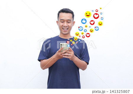 Young man smiles while using smartphone. Colorful emojis float above his head. He enjoys social media interaction. Image captures joyful digital engagement. Perfect for digital marketing visuals 141325807