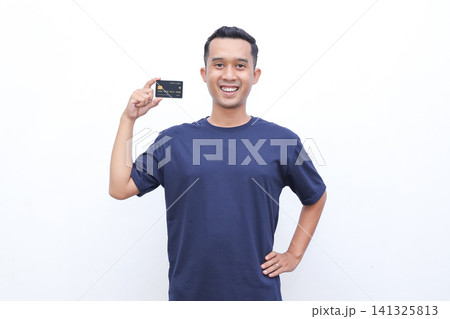 Young man smiles, holding a credit card. He wears a dark blue tshirt. His posture shows confidence and ease. The white background is clean and simple. Ideal for financial, shopping Young man smiles, holding a credit card. He wears a dark blue tshirt. His posture shows confidence and ease. The white background is clean and simple. Ideal for financial, shopping 141325813