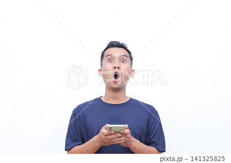 Young man holds phone with wide eyed surprise. He stares ahead, mouth open in astonishment. Simple white background highlights his reaction. Perfect for digital surprises, unexpected news Young man holds phone with wide eyed surprise. He stares ahead, mouth open in astonishment. Simple white background highlights his reaction. Perfect for digital surprises, unexpected news 141325825