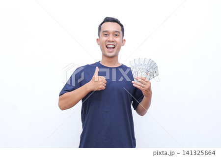 Young man smiles broadly, holding cash and giving a thumbs-up. His expression shows excitement and satisfaction. White background highlights his joyful gesture Young man smiles broadly, holding cash and giving a thumbs-up. His expression shows excitement and satisfaction. White background highlights his joyful gesture 141325864