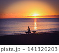 日の出の海辺を歩く人物と犬 141326203