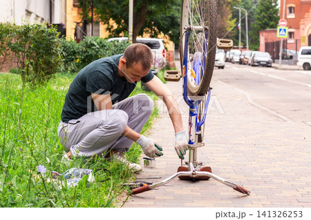 Man fixing bicycle wheel at doorstep. Own vehicle maintenance without service center. Responsible ownership visible. Summer commute switched to pedal power Man fixing bicycle wheel at doorstep. Own vehicle maintenance without service center. Responsible ownership visible. Summer commute switched to pedal power 141326253