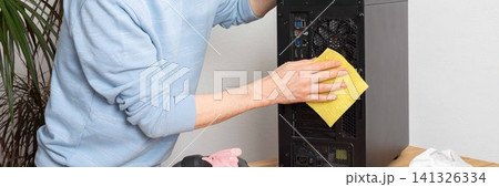A 53-year-old man uses a cloth to clean dust from a computer system unit. He works at a wooden table in a room with plants nearby. Tools are set out for the task, banner A 53-year-old man uses a cloth to clean dust from a computer system unit. He works at a wooden table in a room with plants nearby. Tools are set out for the task, banner 141326334