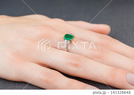 Beautiful female hand with gold ring with diamonds and emerald on gray background Beautiful female hand with gold ring with diamonds and emerald on gray background 141326648