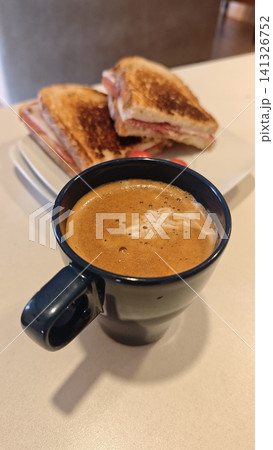 Black coffee served in a ceramic mug on a table, with toasted sandwiches arranged on a plate in the background, creating a casual dining atmosphere 141326752