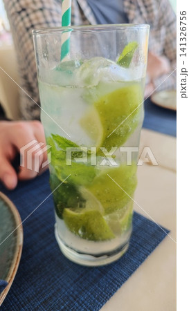 Mojito cocktail featuring fresh mint and lime slices in a tall glass, placed on a blue tablecloth with a hand visible in the background Mojito cocktail featuring fresh mint and lime slices in a tall glass, placed on a blue tablecloth with a hand visible in the background 141326756