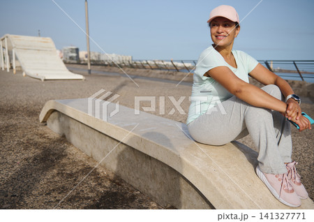 Smiling woman resting after seaside jog in activewear and cap on coastal promenade Smiling woman resting after seaside jog in activewear and cap on coastal promenade 141327771