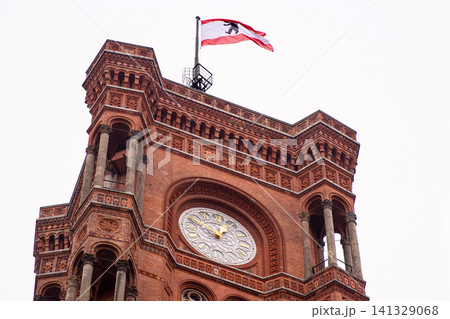 Berlin red town hall with Berlin flag flying in the cloudy sky. Detailed view of Rotes Rathaus clock tower, iconic landmark featuring ornate brickwork representing the city history. Copy space Berlin red town hall with Berlin flag flying in the cloudy sky. Detailed view of Rotes Rathaus clock tower, iconic landmark featuring ornate brickwork representing the city history. Copy space 141329068