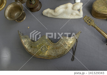 XALO, SPAIN - JUNE 20, 2026.An assortment of brass items and a small marble figure are arranged on gray cloth. The brass items show intricate designs. The marble figure appears to be reclining and add XALO, SPAIN - JUNE 20, 2026.An assortment of brass items and a small marble figure are arranged on gray cloth. The brass items show intricate designs. The marble figure appears to be reclining and add 141329343