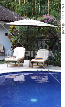 Poolside lounge chairs beneath large patio umbrella beside blue tiled swimming pool in tropical Bali garden with bamboo, bougainvillea, stone wall, and tranquil resort setting Poolside lounge chairs beneath large patio umbrella beside blue tiled swimming pool in tropical Bali garden with bamboo, bougainvillea, stone wall, and tranquil resort setting 141331971