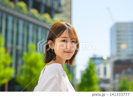 笑顔の女性、都会の街並みで新緑を背景に立つ 笑顔の女性、都会の街並みで新緑を背景に立つ 141333777