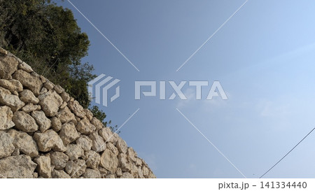 A horizontal wide angle perspective captures a curved dry stone wall built from weathered limestone blocks below a clear light blue sky. Green tree foliage emerges from behind the stony structure, 141334440
