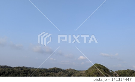 A horizontal wide angle perspective captures lush green forest hills stretching across the horizon under a vast blue sky. Scattered white clouds drift gently above the rolling tropical mountain 141334447