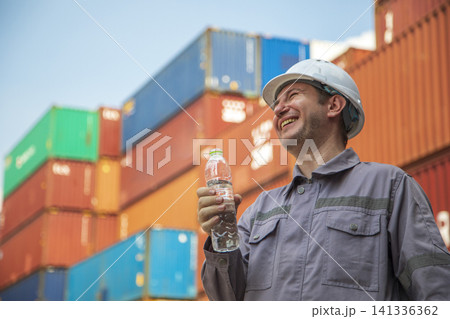 Worker man drinks water from a plastic bottle at container ship yard. Worker man drinks water from a plastic bottle at container ship yard. 141336362