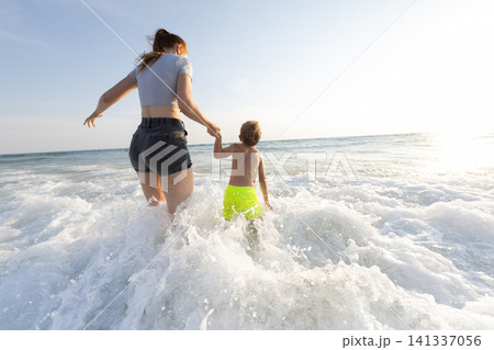 Woman and child running into ocean waves during sunset, both wearing summer clothing, with the sun reflecting on the water surface in a coastal setting Woman and child running into ocean waves during sunset, both wearing summer clothing, with the sun reflecting on the water surface in a coastal setting 141337056