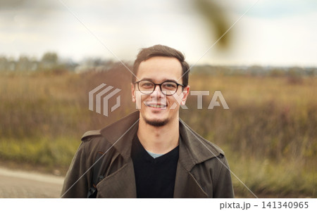 Portrait of handsome young man with glasses smiling outdoors in autumn field. Happy confident caucasian male looking at camera wearing stylish coat, autumn season concept Portrait of handsome young man with glasses smiling outdoors in autumn field. Happy confident caucasian male looking at camera wearing stylish coat, autumn season concept 141340965