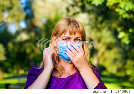 Middle-aged woman putting on protective medical face mask outdoors in park. Mature caucasian female wearing blue disposable mask, concept of allergy, virus prevention, air pollution and healthcare Middle-aged woman putting on protective medical face mask outdoors in park. Mature caucasian female wearing blue disposable mask, concept of allergy, virus prevention, air pollution and healthcare 141340968