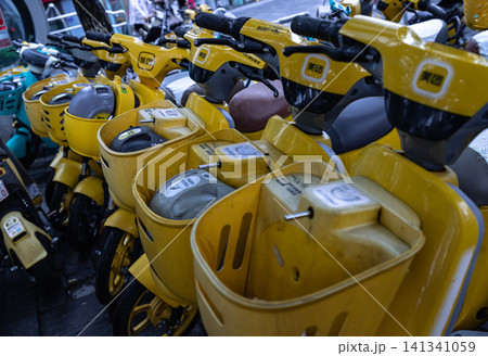 A perspective close-up shot of vibrant yellow electric rental scooters with front baskets neatly lined up on an urban city sidewalk. A perspective close-up shot of vibrant yellow electric rental scooters with front baskets neatly lined up on an urban city sidewalk. 141341059