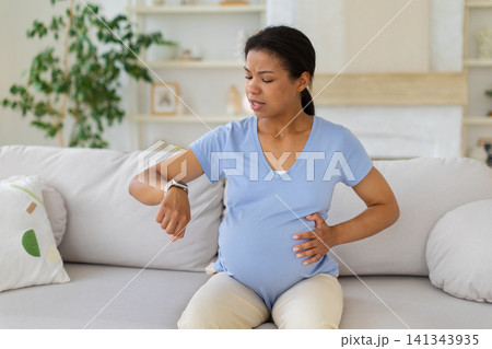Black pregnant woman checking her smartwatch while holding her belly on sofa Black pregnant woman checking her smartwatch while holding her belly on sofa 141343935