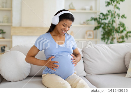Happy Black pregnant woman wearing headphones smiling down and cradling her belly 141343936