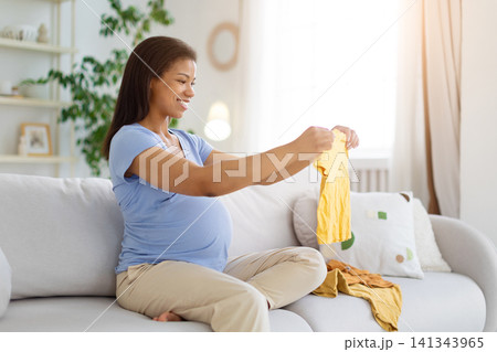 Black pregnant woman sitting on sofa and holding up yellow baby garment Black pregnant woman sitting on sofa and holding up yellow baby garment 141343965