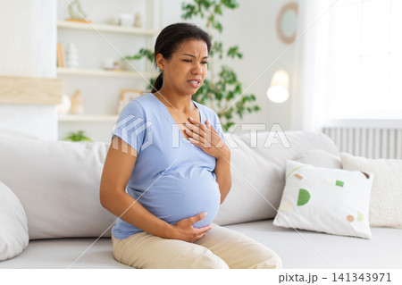 Black pregnant woman sitting on sofa touching her chest and belly Black pregnant woman sitting on sofa touching her chest and belly 141343971