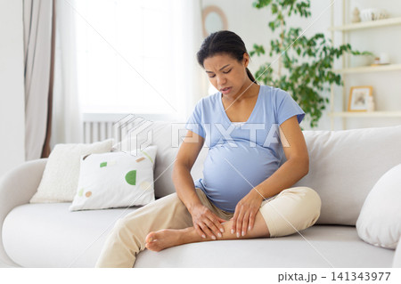 Black pregnant woman sitting on sofa and massaging ankle, showing leg pain Black pregnant woman sitting on sofa and massaging ankle, showing leg pain 141343977