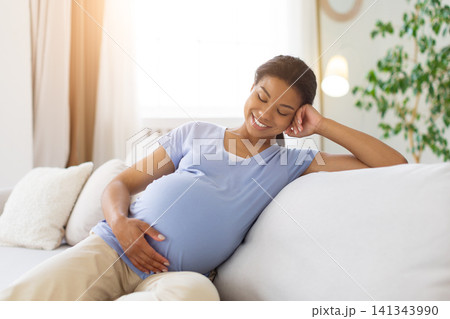 Black pregnant woman resting on sofa with eyes closed, touching her belly Black pregnant woman resting on sofa with eyes closed, touching her belly 141343990