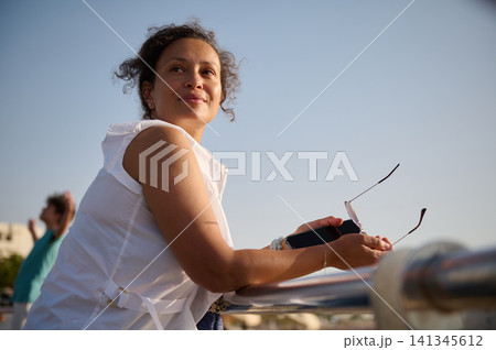 Smiling woman leaning on railing outdoors holding sunglasses and phone on a sunny summer day 141345612