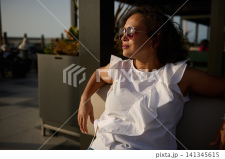 Woman relaxing on outdoor patio at sunset wearing sunglasses and white ruffled top 141345615