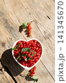 Heart shaped bowl filled with fresh red currants on weathered wooden table, rustic summer still life with natural light, soft shadows, green sprigs, glossy fruit and warm seasonal color 141345670