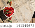 Heart-shaped white bowl filled with fresh red currants on weathered wooden table, rustic tabletop scene with loose green leaves, bright sunlight, natural food styling and seasonal freshness 141345672