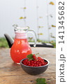Glass pitcher of pink currant drink beside ceramic bowl of fresh red currants on weathered wooden table in garden setting with soft natural light and rustic outdoor atmosphere 141345682