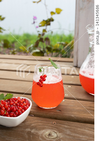 Bright red currant drink in clear glass with berry garnish on rustic wooden table, served beside fresh currants, bowl, pitcher, natural outdoor summer setting, with red, currant, drink, fruit detail Bright red currant drink in clear glass with berry garnish on rustic wooden table, served beside fresh currants, bowl, pitcher, natural outdoor summer setting, with red, currant, drink, fruit detail 141345686
