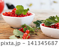 Red currants piled in white ceramic bowl with glossy green leaf, scattered sprigs on rustic wooden table in soft natural light, vivid fresh summer fruit still life closeup 141345687