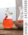 Glass of red currant drink with berry sprig, ice condensation, on light wooden table, bright outdoor summer setting with blurred jug, fresh foliage and natural refreshment 141345688