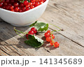 Close view of bright red currants spilling from white bowl onto weathered wooden table, with attached green leaves, rustic texture, soft sunlight, glossy fruit, fresh harvest feel 141345689