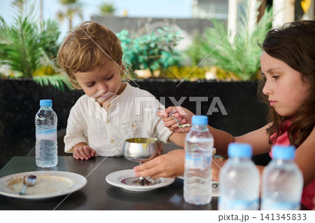 Young siblings sharing dessert at outdoor cafe as sister feeds little brother Young siblings sharing dessert at outdoor cafe as sister feeds little brother 141345833