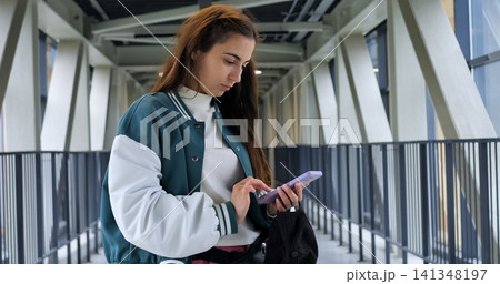 Young woman offering friendly glance in pedestrian corridor, slight smile while holding phone and bag, confident posture with relaxed shoulders, modern geometric background and soft daylight creating 141348197