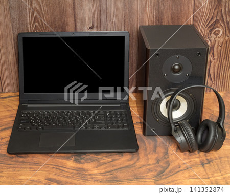 open black laptop stands on a wooden table next to an audio speaker and headphones 141352874