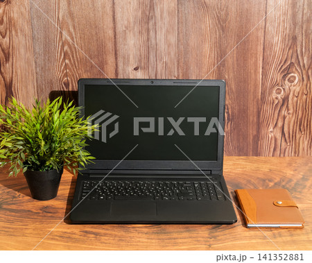 open laptop stands on a table between a green plant in a pot and a leather notebook open laptop stands on a table between a green plant in a pot and a leather notebook 141352881