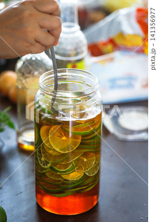 Hand stirring fresh lime slices in a glass jar filled with honey during homemade honey lime infusion preparation. Healthy drink, natural ingredients, and food preservation concept 141356977