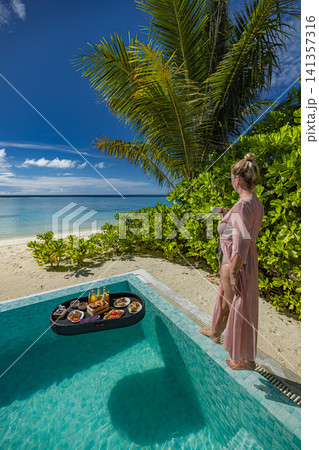Woman standing at private pool edge with floating breakfast under palm trees. Guest in villa garden enjoying tropical morning, lush greenery and sunny sky. Aspirational resort lifestyle scene Woman standing at private pool edge with floating breakfast under palm trees. Guest in villa garden enjoying tropical morning, lush greenery and sunny sky. Aspirational resort lifestyle scene 141357316