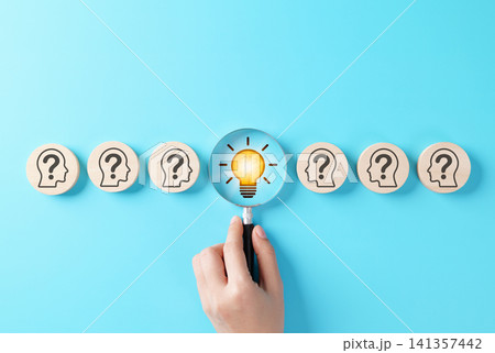 Creative thinking and problem-solving concept. Hand holding magnifying glass discovering a light bulb among wooden icons of question marks on heads, Symbol of idea, innovation and finding solutions. Creative thinking and problem-solving concept. Hand holding magnifying glass discovering a light bulb among wooden icons of question marks on heads, Symbol of idea, innovation and finding solutions. 141357442
