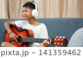 Happy Asian boy wearing headphones playing acoustic guitar on sofa at home. 141359462