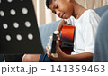 Happy Asian boy smiling while playing acoustic guitar and practicing at home. Cute young child learning to play musical instrument looking at music sheet stand in living room. 141359463