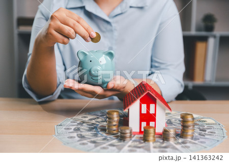 Home savings concept with blue piggy bank, coin stacks, cash, and red house model on office desk for mortgage deposit, down payment, property investment, financial security, and future ownership. 141363242